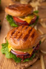 Hot juicy burger on a wooden cutting board. Cheeseburger with beef, cheese, vegetables, crispy buns and salt fries on a rustic table. Fresh meat. Tasty meal.