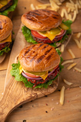 Fresh hot burger with french fries. Meal on a cutting kitchen board on a wooden table. Cheeseburger with meat, cheese, tomatoes, cucumbers and onions. The concept of street junk food. Vertical picture