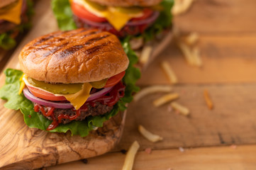 Fresh hot juicy burger with salty french fries. Meal on a cutting kitchen board on a wooden table. Cheeseburger with meat, cheese, tomatoes, cucumbers and onions. American cuisine. Rustic style