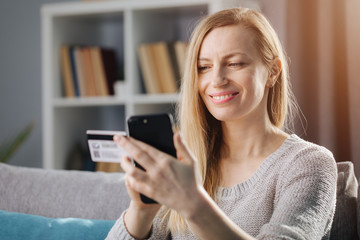 Fototapeta na wymiar Attractive mature woman with long blond hair doing online shopping while sitting at home. Smiling lady in casual clothing using credit card and smartphone for payment.