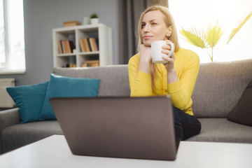 Charming mature lady with long hair sitting on couch in front of opened laptop with cup of coffee in hands. Thoughtful woman in casual outfit having break during working day at home.