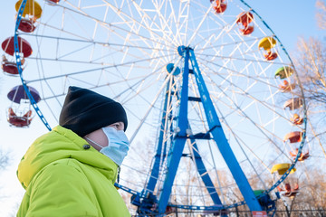 a child in a hospital anti virus mask and an autumn jacket in a city amusement Park looks at the Ferris wheel with sadness in his eyes