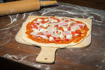 Pizza roh auf Holzschieber mit Tomatensauce, Mozzarella, Kochschinken, prosciutto cotto, Tomaten,...