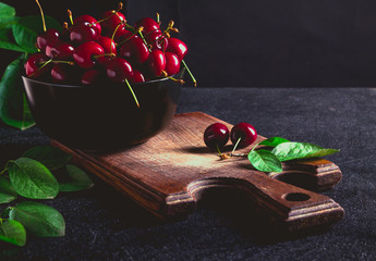 Ripe sweet cherries with green leaves in a black bowl on a brown cutting board on a black background