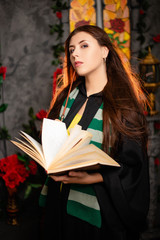 A young woman in a black mantle with a striped scarf around her neck casts a spell from a book and...