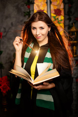 A young woman in a black mantle with a striped scarf around her neck casts a spell from a book and...