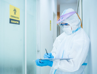 Asian doctor in personal protective suit wearing surgical mask and writing on quarantine patient chart, work hard without depression in covid-19 crisis. Medical and coronavirus concept.