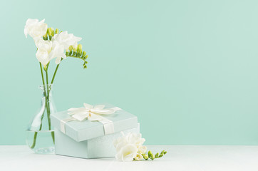 Fresh delicate white flowers freesia and festive ajar square gift box with ribbon and knot in green mint menthe interior on white wood board.