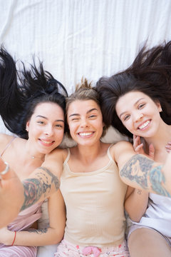 Close up of smiling group of girls in pajamas having fun at home in bedroom. Three pretty girls taking selfie after shower in bedroom. Concept of friendship, skincare. Top view
