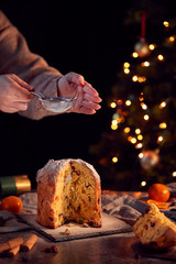 Hand Shaking Icing Sugar Over Christmas Panettone On Table Set For Meal With Tree Lights Behind - obrazy, fototapety, plakaty