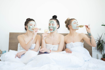 Obraz na płótnie Canvas Three funny happy beautiful girlfriends making face clay masks, laughing and eating cucumbers. Stay at home concept. Skin care and treatment, spa, natural beauty and cosmetology concept.