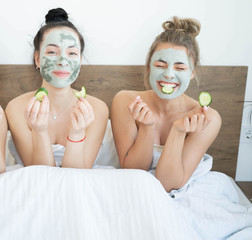 Two funny happy beautiful girlfriends making face clay masks, laughing and eating cucumbers. Stay at home concept.