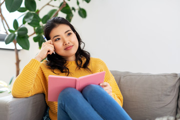 people and leisure concept - thinking asian young woman with diary or notebook sitting on sofa at home