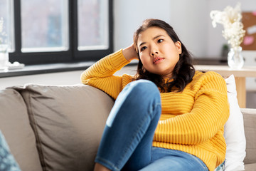 people and leisure concept - worried asian young woman in yellow sweater sitting on sofa at home