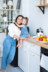 happy mom and daughter about in the kitchen. mom brunette in a white shirt and jeans, daughter in denim overalls. kitchen in a Scandinavian style. beautiful family.fruit plate in the background