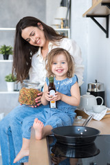happy mom and daughter are cooking in the kitchen. mom brunette in white shirt and jeans, daughter in denim overalls. bright and spacious kitchen in Scandinavian style. a plate of fruit on the table
