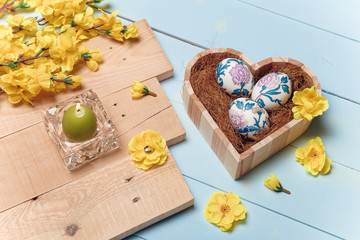 Close up shot of Heart shaped box with decorated Easter eggs, yellow flowers, burning candle and brown boards on wooden blue background. Spring and Easter concept.