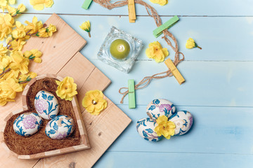 Heart shaped box with decorated Easter eggs, yellow flowers, burning candle and brown boards on wooden blue background. Spring and Easter concept. Top view, flatlay, copy space.