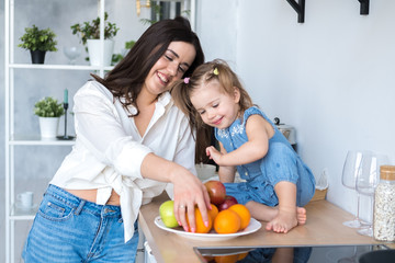 happy mom and daughter eating fruit in the kitchen. a plate with apples, oranges, pears and lemon stands on the table. bright and spacious kitchen in Scandinavian style. healthy food for children