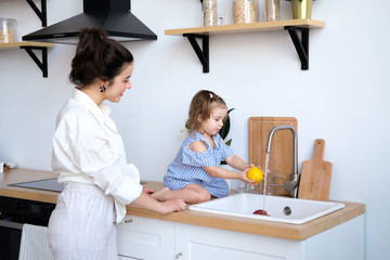 A beautiful young woman with her two year old daughter is washing fruit in the kitchen sink. Fresh bright fruits, apples, oranges, pears. Healthy food for children. The baby helps her mother.