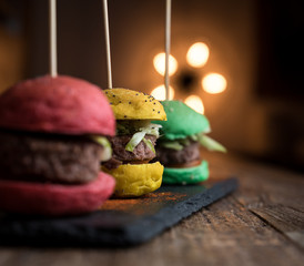 Mini hamburger colorati di manzo su piatto di ardesia, tavolo in legno e background con luce calda