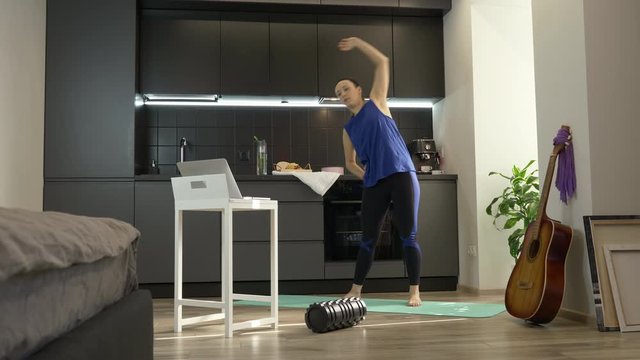 Young woman stretches in kitchen at home. Sporty fit female in athletic workout clothes doing stretching exercises at home using online fitness app on laptop computer. Girl doing morning exercises