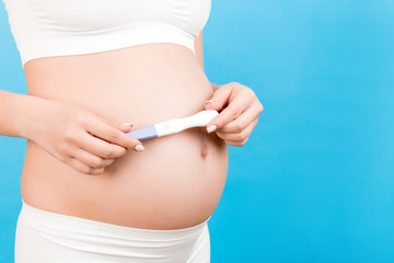 Close up portrait of pregnant woman in white underwear holding positive pregnancy test against her belly at blue background. Healthy pregnancy. Copy space