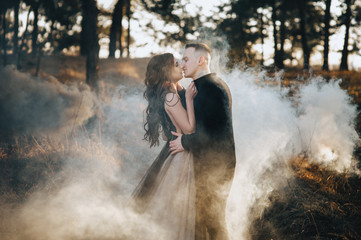 Fototapeta na wymiar A loving bridegroom in a black cardigan and a cute bride in an expensive dress are holding a pyrotechnic device with smoke in their hands and hugging. Wedding portrait of the newlyweds.