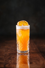 delicious orange sour cocktail in the long etched glass with ice and orange slice, dark wooden background, vertical, side view