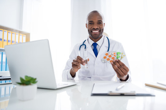 Photo of family doc dark skin cheerful guy assistant hold drugs variety prescription water glass help control risk epidemic disease prophylactics wear white lab coat office clinic indoors
