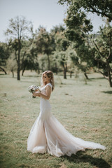 Fototapeta na wymiar Portrait of a wedding bride walking in a white modern hipster dress. Caucasian blonde bride in a vintage dress in the forest on sunset. Attractive lady posing with a bouquet of flowers