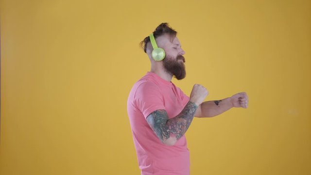 Close-up of tattooed man caucasian with beard and moustache in pink t-shirt listening to music hands up,and enjoying on headphones, isolated on yellow background