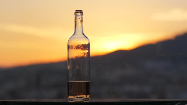 Male hand take glass and make sip of weak wine, then put in back. Open bottle stand near, blurred sunset sky seen on background. Man relaxing at evening time