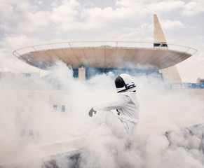 An astronaut in a spacesuit stands in a cloud of smoke against the backdrop of urban structures and architecture. 