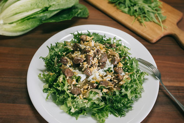 Delicious warm salad with green leaves of frisee, chicken hearts, meat, egg and cheese. Served on a white plate and ready to eat. Cooking at home concept