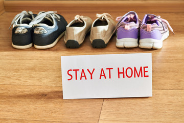 Family shoes are standing on the floor near the threshold with the inscription on paper STAY AT HOME. Stay safe by stay home medicine concept. Protection campaign or measure from coronavirus, COVID-19