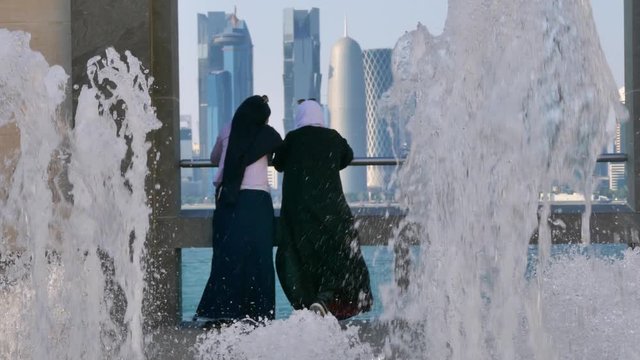 Rear shot of two Arab girls with Islamic hijab looking at skyscrapers behind fountains