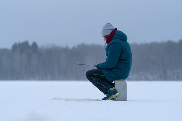 Fototapeta na wymiar A man on a frozen snowy lake. Side view. Winter fishing. Fishing rod in the hands. Ice hole. White snow. Snow-covered forest and lake shore on the horizon. Grey sky.