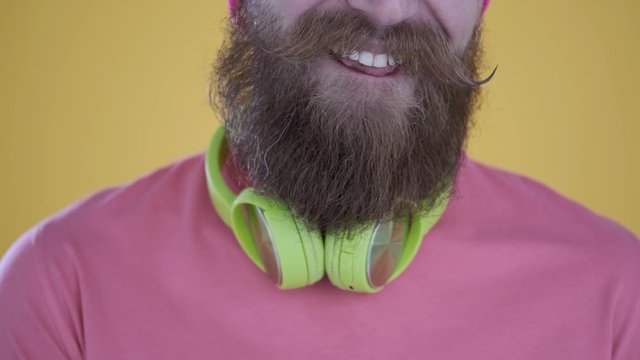 Close-up of tattooed Caucasian man with beard and mustache in pink t-shirt and hat eating a Chupa Chups,sweet candy,looking at camera,with headphones isolated on yellow background