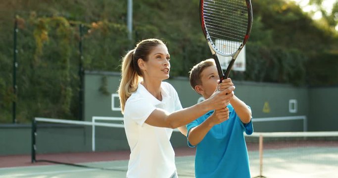 Caucasian young beautiful woman teaching her son how to hit ball with racket on sunny summer day. Pretty female instructor showing to boy how to play tennis on sport court.