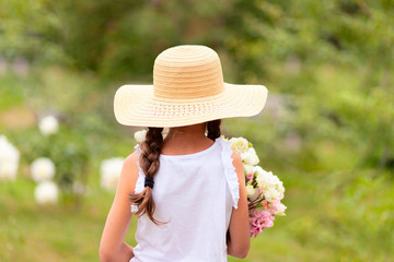 A girl with pigtails holds in her hands a bouquet of white and pink peonies. A child in a wide-brimmed straw hat on a sunny summer day. Green background. Back view.