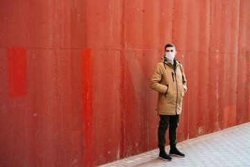 a young man in a brown jacket with a medical mask on his face, fighting against the corona 19 virus infection on the street, near the red wall.