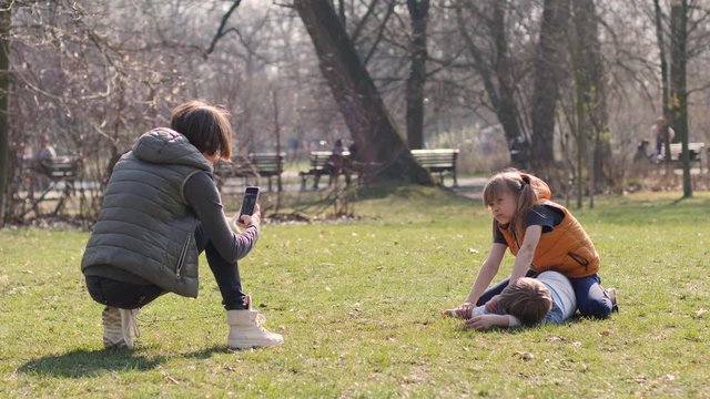Young mother takes phone picture of her children daughter and son lying on park grass family having fun together outdoors