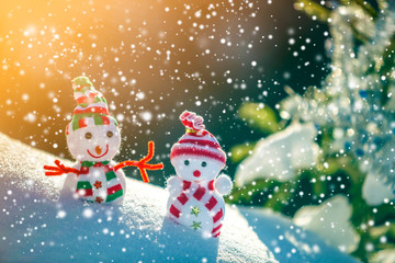 Two small funny toys baby snowman in knitted hats and scarves in deep snow outdoors near pine tree branch. Happy New Year and Merry Christmas greeting card.