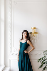 Brunette girl in a long green dress stands near a light wall