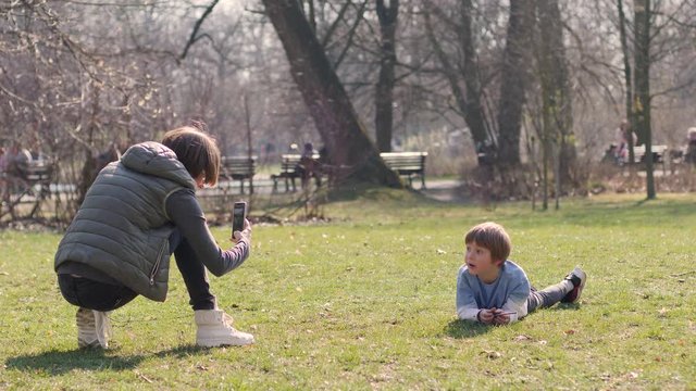 Young mother takes phone picture of her son kid lying on park grass family having fun together outdoors