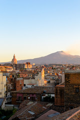 Fototapeta premium Catania, Sicily in Italy. Aerial view of the city roofs at sunset with the incredible Etna vulcano smoking in the background, nice warm colors and soft light.