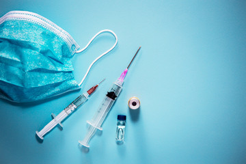 Medical items flat laid on top of light blue background, with available copy space. Surgical face mask, vaccine syringes with needles and medicine bottles.