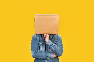 Hombre con caja de cartón en la cabeza con gesto pensativo y mano en la barbilla sobre fondo amarillo liso brillante aislado. Vista de frente. Copy space - obrazy, fototapety, plakaty
