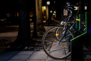 Black bicycle parked at night with city park road in the background. Bicycle parking rack. Smart, automatic, electric rack with built in lock system. E-Bike concept. Close up, copy space